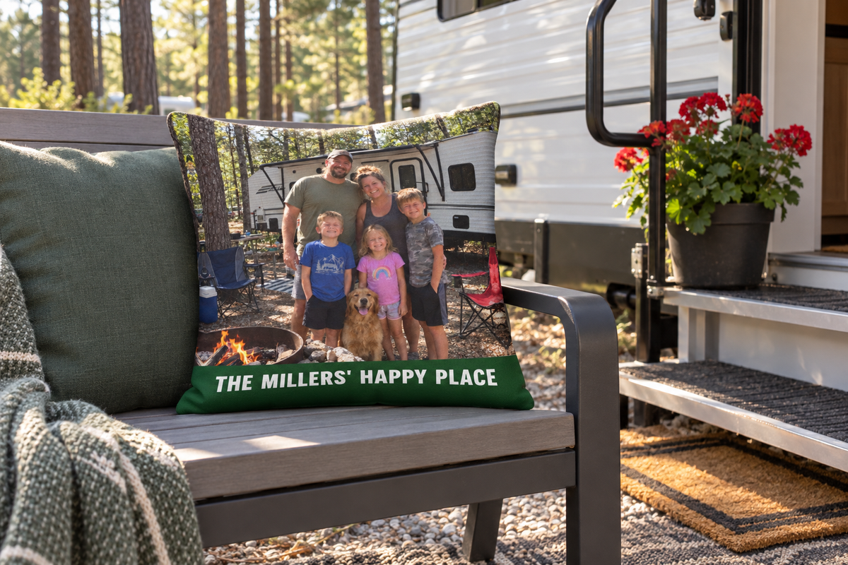 Outdoor pillow with a family camper photo and The Millers' Happy Place on a bench by a travel trailer door