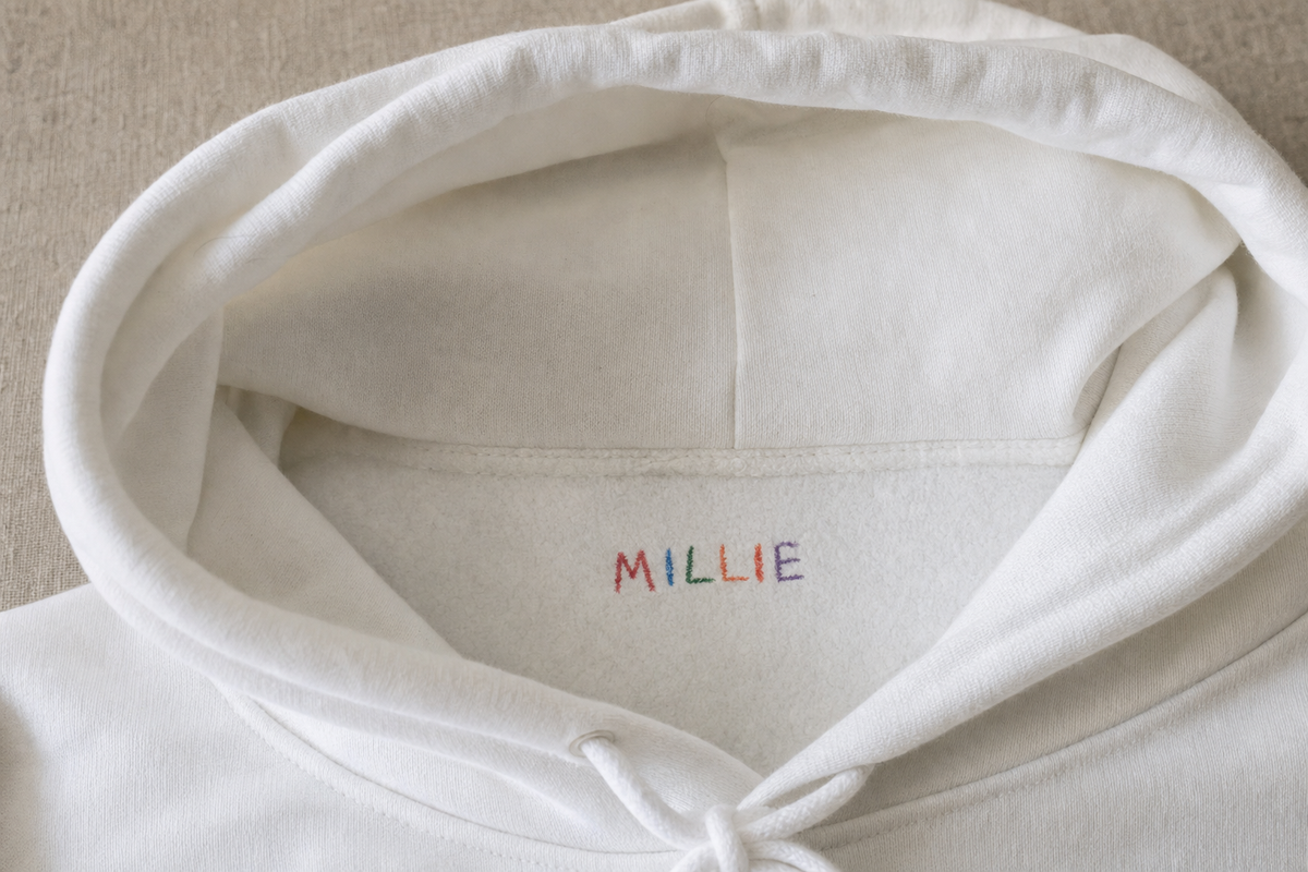 A child's crayon signature printed at the inside neck of a white custom hoodie where a garment tag would sit
