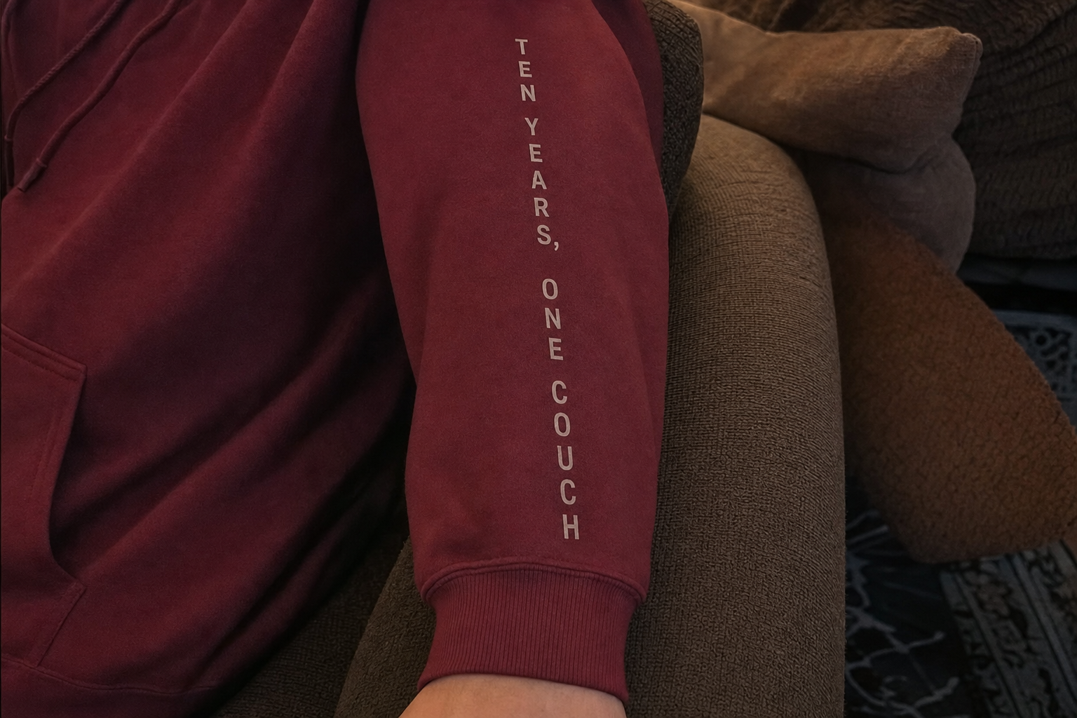 Thin uppercase text reading Ten Years One Couch printed down the sleeve of a maroon custom hoodie with the ribbed cuff visible