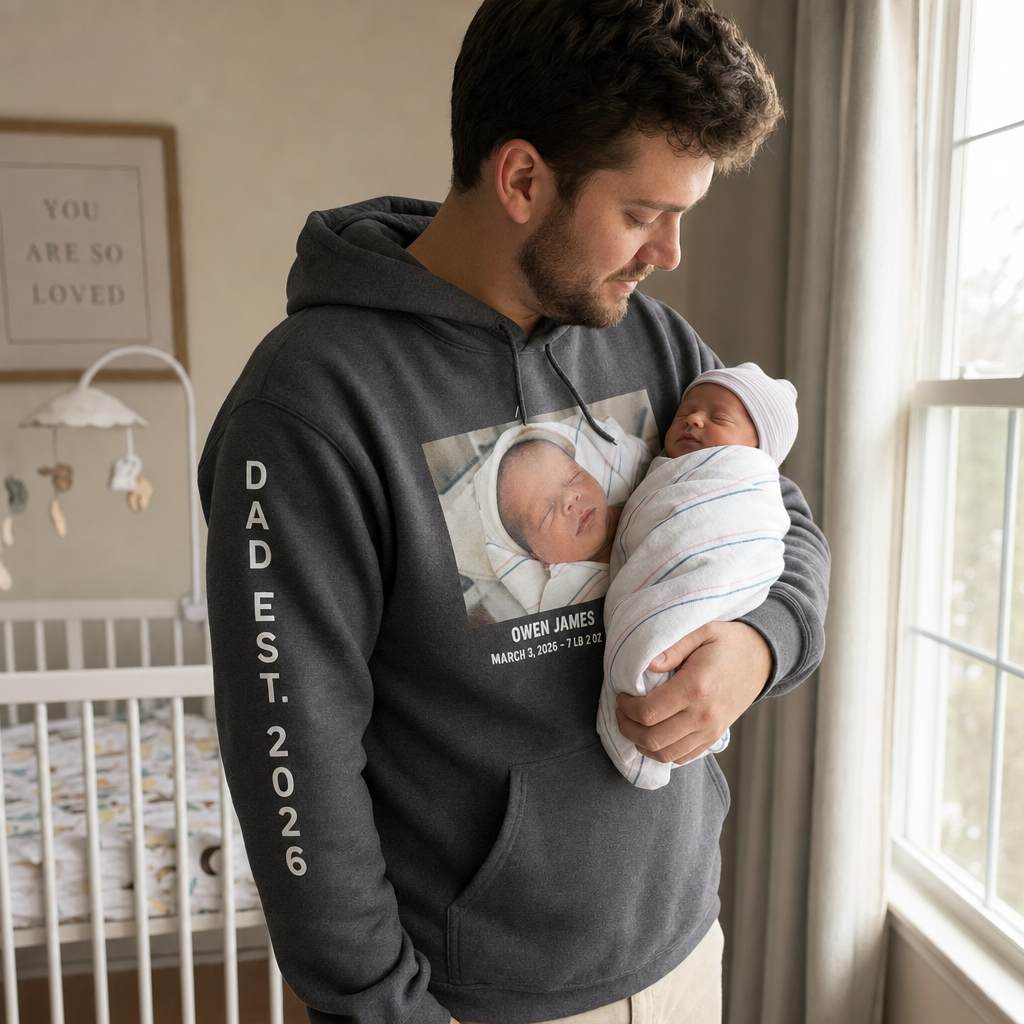 A new dad in a dark heather custom hoodie with Dad Est 2026 printed down the sleeve, rocking a newborn by a window