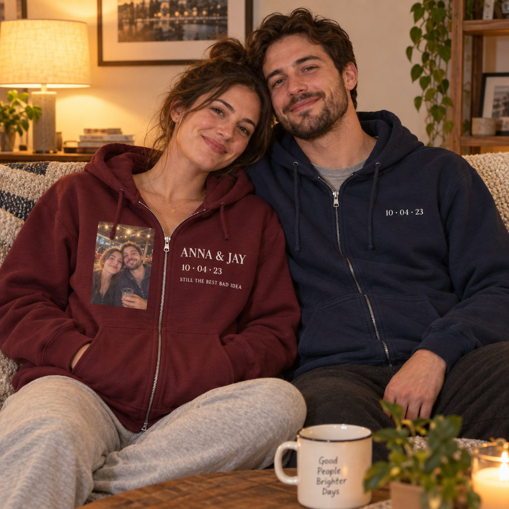 A couple on a couch in custom zip-up hoodies, hers maroon with a photo of the two of them on one side of the zipper and their names and date on the other